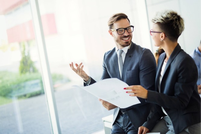 Zwei Personen in formeller Geschäftskleidung sitzen in einem modernen Büro mit großen Glasfenstern. Eine Person hält ein Dokument in der Hand und unterhält sich. Natürliches Licht und Grün im Hintergrund schaffen eine professionelle und zugleich entspannte Atmosphäre.