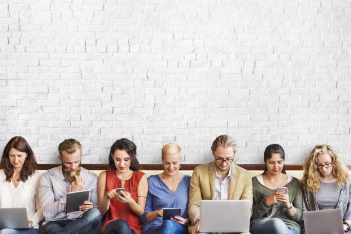 Eine vielfältige Gruppe von Personen sitzt vor einer weißen Ziegelwand, jede:r beschäftigt sich mit verschiedenen digitalen Geräten wie Laptops, Tablets und Smartphones. Die Szenerie wirkt locker und modern, mit Fokus auf Technologie-Nutzung. Das Bild vermittelt Vernetzung und Interaktion in einer neutralen, hellen Umgebung.