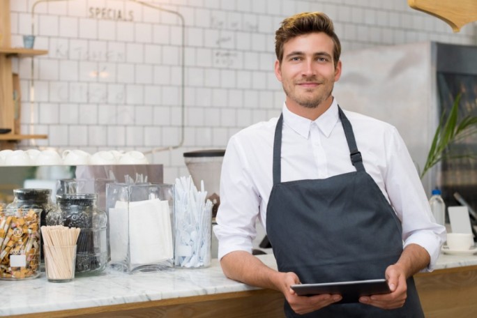 A cafe worker wearing a white shirt and dark apron stands at a counter holding a tablet. The counter features jars of snacks, napkins, and utensils, with a tiled wall and menu board in the background. The setting suggests a casual and modern cafe environment.