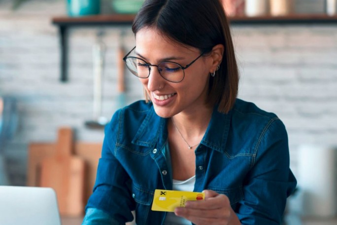 A woman is seated in a modern kitchen holding a bright yellow card with visible text and symbols. The background features shelves with jars and kitchen utensils, creating a casual and homey atmosphere. She is wearing a denim shirt, accessorized with earrings, and appears to be engaged in an activity involving the card.