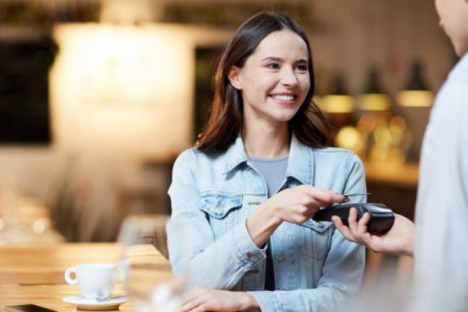 A woman in a casual denim jacket is seen making a contactless payment using a card reader. The setting is a cozy café with warm lighting and wooden tables. A white coffee cup is visible on the table, adding to the relaxed atmosphere.