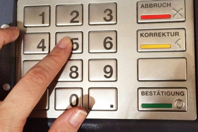A close-up image of a hand pressing the number 5 on a metallic keypad, likely part of an ATM or payment terminal. The keypad includes numeric buttons from 0 to 9 and labeled function keys such as 'Abbruch', 'Korrektur', and 'Bestätigung'. The setting suggests a financial or transactional activity.