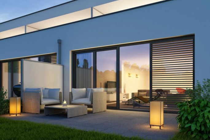 A contemporary patio featuring wicker outdoor furniture and soft lighting at dusk. The setting includes a sleek modern building with large glass doors and horizontal blinds. Warm ambient lighting creates a cozy atmosphere, complemented by greenery in the foreground.