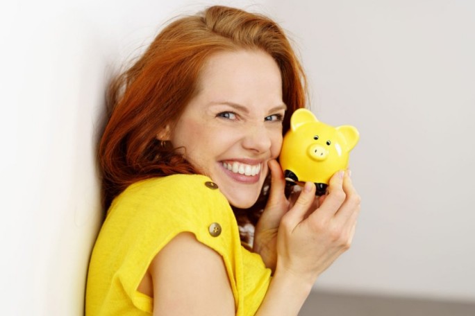 A person wearing a bright yellow outfit holds a small yellow piggy bank. The setting appears to be indoors with a neutral white background. The piggy bank features a cheerful design with black accents on its base.