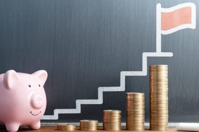 A pink piggy bank sits next to stacks of gold coins arranged in ascending order, symbolizing financial growth. A chalk-drawn staircase leads to a red flag on a gray background, emphasizing goals and savings. The image conveys themes of investment, progress, and achievement.