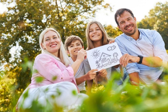 A group of people sitting outdoors in a lush green park, holding a hand-drawn sketch of a house surrounded by trees and a sun. The setting features bright natural light and a relaxed atmosphere. The drawing is simple and black-and-white, emphasizing creativity and family bonding.