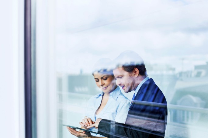 Zwei Personen interagieren im Freien mit einem Tablet. Die Szene ist durch eine Glasscheibe zu sehen. Im Hintergrund ist eine moderne Stadtkulisse mit sanftem Licht und Spiegelungen zu erkennen. Das Bild vermittelt eine professionelle und kooperative Atmosphäre.