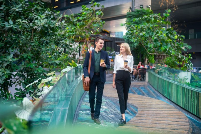 Zwei Personen in Businesskleidung spazieren auf einem Holzsteg durch einen üppigen Indoor-Garten. Der Garten besticht durch viel Grün, Glasgeländer und eine moderne Architektur. Beide halten Coffee-to-go-Becher in der Hand, was auf ein lockeres Gespräch oder eine kurze Pause hindeutet. Die Atmosphäre ist angenehm mit einer Mischung aus natürlichem und künstlichem Licht beleuchtet.