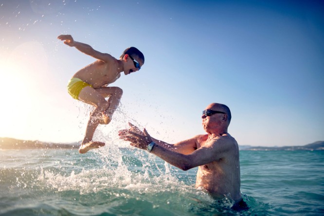 Ein Kind in gelber Badehose springt ins Wasser, ein Erwachsener fängt es auf. Die Szene spielt sich an einem sonnigen, offenen Meer mit strahlend blauem Himmel und sanften Wellen ab. Der Erwachsene trägt eine Sonnenbrille, was die sommerliche Stimmung des Bildes unterstreicht.