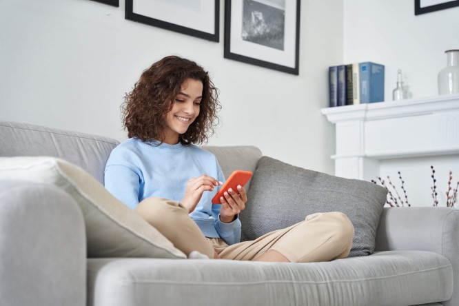 Eine Frau sitzt auf einem grauen Sofa in einem modernen Wohnzimmer und benutzt ein rotes Smartphone. Die Einrichtung ist gemütlich und minimalistisch gehalten, mit gerahmten Bildern, Büchern und einem weißen Kaminsims im Hintergrund. Sie trägt einen hellblauen Pullover und eine beige Hose, was eine entspannte und behagliche Atmosphäre schafft.