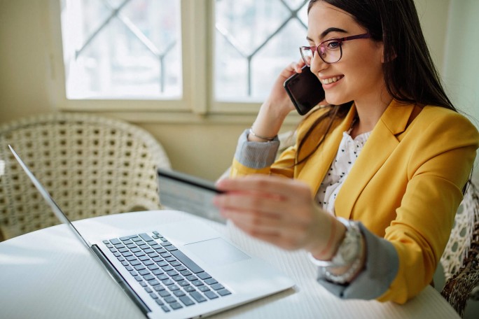 Eine Frau in einem gelben Blazer sitzt an einem Tisch, hält eine Kreditkarte in der Hand und telefoniert. Vor ihr steht ein geöffneter Laptop, was auf Online-Shopping oder eine Finanztransaktion hindeutet. Der Raum wirkt hell und freundlich, mit natürlichem Licht, das durch die Fenster fällt.