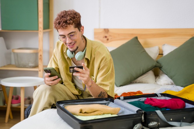 Young man sitting on bed with suitcase open and using smartphone and credit card for online booking of hotel room